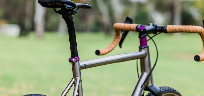 Partial view of a custom-built Bossi Grit SX titanium bicycle with tan handlebar tape & purple accents, in a park setting