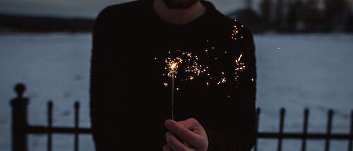 A man holding a sparkler. He is wearing a black jumper, a body of water behind him.