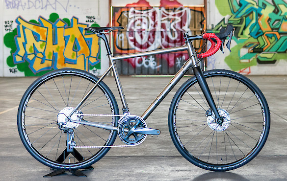 A custom-built titanium Bossi Grit gravel bike with bright red handlebar tape, shot inside a colourful graffitied car park
