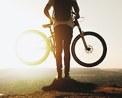 A mountain biker, facing away from the camera, holding up a mountain bike at waist height and looking over the horizon.