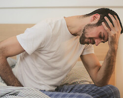 A man sitting on the side of an unmade bed, holding his head in apparent pain.