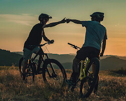 Two mountain bikers standing on a hill, bumping fists as the sun sets in the distance.