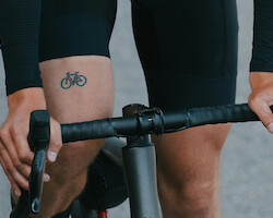 Close-up of the bicycle tattoo on a bike rider's thigh. They are wearing black lycra and riding a drop-bar bike.