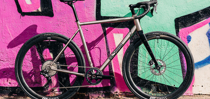 Titanium gravel Bossi Grit bicycle, custom-built by Bio-Mechanics Cycles & Repairs, photographed against a colourful pink and green mural.